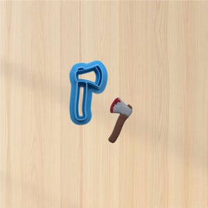 May include: A blue plastic axe-shaped cookie cutter next to a small axe-shaped object with a brown handle, gray blade, and red accents. The objects are on a light wood-grain surface.