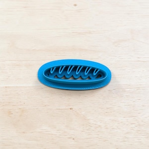 Könnte beinhalten: Ein hellblauer, ovaler Ausstecher mit einem wellenförmigen Innendesign. Der Ausstecher besteht aus Kunststoff und liegt auf einer hellen Holzoberfläche. Das Design zeigt eine Reihe von vertikalen Bögen über einer wellenförmigen Linie.
