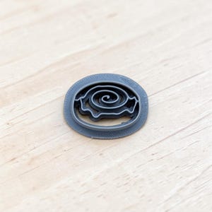 May include: A dark gray, oval-shaped cookie cutter featuring an intricate, central spiral design. The object has a flat, wide outer rim and a raised, thin inner wall that forms the spiral pattern, constructed from a solid, matte-finished material.