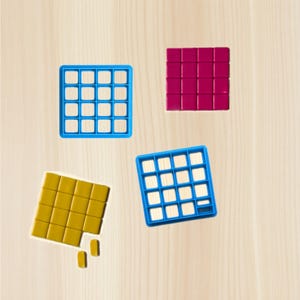 May include: Four colourful square-shaped objects are arranged on a light wood surface. A blue grid, a magenta grid, a yellow grid, and a second blue grid are visible. The yellow grid has two detached pieces.