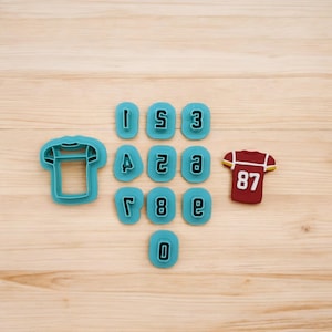 May include: A set of blue plastic cookie cutters shaped like football jerseys and numbers 0-9. The numbers are in a block font style. There is a red cookie in the shape of a football jersey with the number 87 on it.