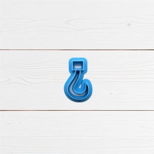 May include: A blue plastic question mark-shaped cookie cutter. The cutter has a square top and a curved, hook-like bottom. The object is set against a white wooden background.