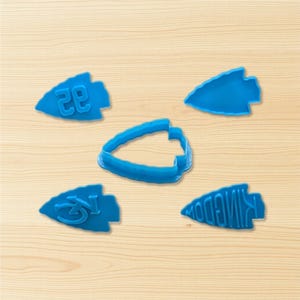 May include: A collection of five blue plastic arrowhead-shaped cookie cutters. One cutter has the number "28", another has the letters "KC", and a third has the word "KINGDOM". The cutters are arranged on a light wood surface.