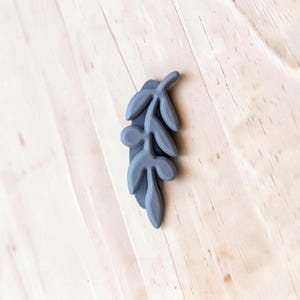 May include: A matte gray, three-dimensional leaf-shaped object featuring a central stem with four symmetrical, rounded leaf segments extending outward. The surface displays a subtle, fine-lined texture consistent with additive manufacturing.