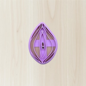 May include: A purple, teardrop-shaped cookie cutter with a layered design. The cutter has a cross-like structure in the center and is set against a light wood grain background. The design is intricate and detailed.