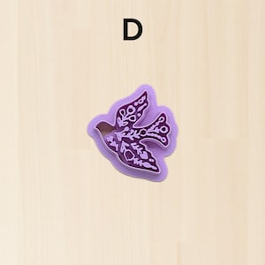Puede incluir: Un cortador de galletas morado en forma de paloma con intrincados dise&ntilde;os florales. La letra "D" est&aacute; impresa en negro en la parte superior. El cortador es de pl&aacute;stico y est&aacute; sobre un fondo claro de vetas de madera.