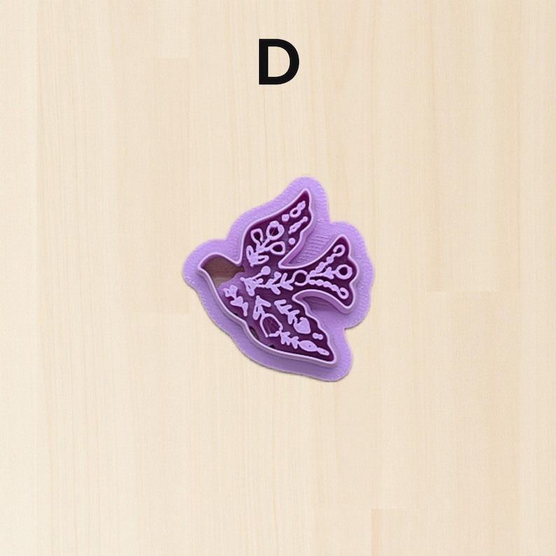 Puede incluir: Un cortador de galletas morado en forma de paloma con intrincados dise&ntilde;os florales. La letra "D" est&aacute; impresa en negro en la parte superior. El cortador es de pl&aacute;stico y est&aacute; sobre un fondo claro de vetas de madera.