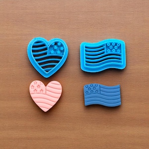 May include: Four cookie cutters in the shape of the American flag and a heart. Two are blue, and two are pink. The heart and flag designs include stars and stripes, perfect for patriotic baking.