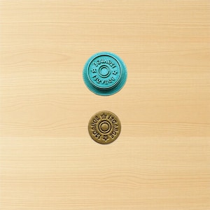 May include: Two round, embossed objects on a light wood surface. One is teal with the text "12 GAUGE" and the other is brown with the text "12 GAUGE". Both have a central circle and star-like designs.