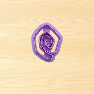 May include: A purple plastic cookie cutter in the shape of a stylized rose. The cutter has a hexagonal outer shape with a spiral design in the center. The cutter is on a light wood-grain surface.