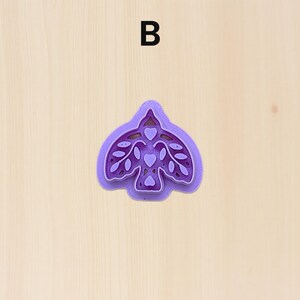 Puede incluir: Un cortador de galletas morado con forma de p&aacute;jaro con detalles de hojas y corazones. La letra "B" est&aacute; impresa en negro en la parte superior. El cortador est&aacute; sobre una superficie de madera clara.