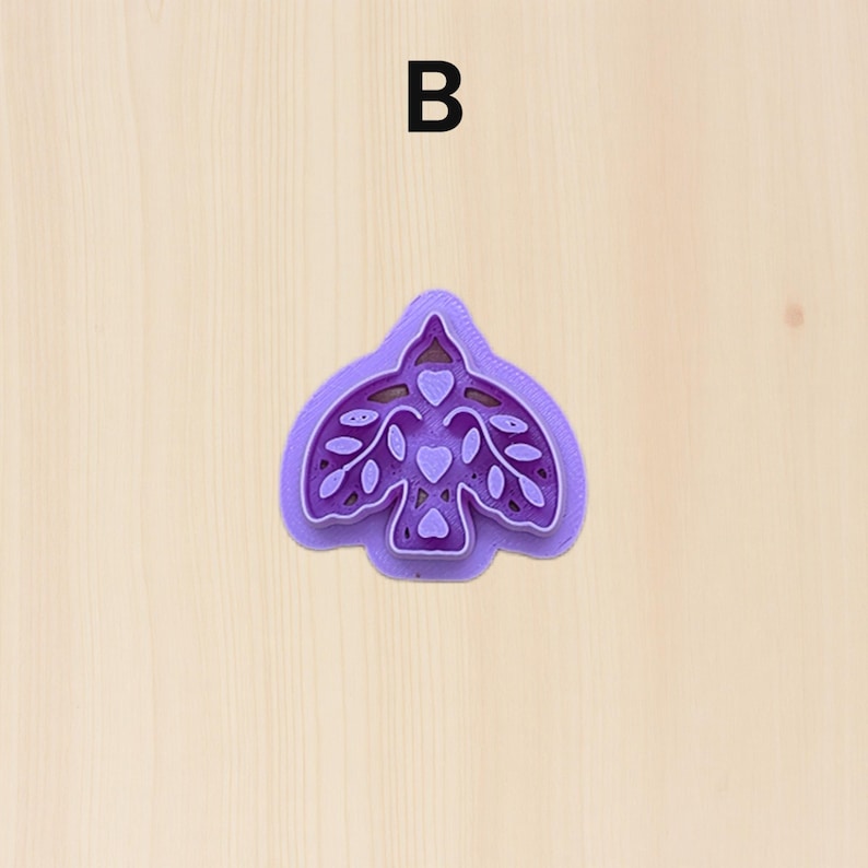 Puede incluir: Un cortador de galletas morado con forma de p&aacute;jaro con detalles de hojas y corazones. La letra "B" est&aacute; impresa en negro en la parte superior. El cortador est&aacute; sobre una superficie de madera clara.