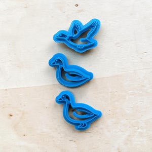 May include: Three blue plastic duck-shaped cookie cutters. One duck is in flight, and the other two are in a resting position. The cutters are on a light-colored wooden surface. These cutters are ideal for baking.