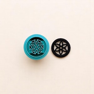 May include: A teal blue plastic cookie cutter with a black plastic stencil of a geometric flower design.