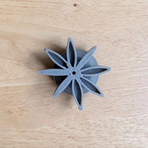 May include: A gray, star-shaped cookie cutter with eight pointed petals radiating from a central circular base. The object features a distinct 3D-printed texture with visible layer lines on the surface of the petals and the cylindrical base.