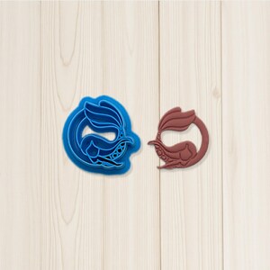 Pode incluir: Um cortador de biscoitos azul em forma de sereia ao lado de um biscoito em forma de sereia marrom. Os cortadores de biscoitos estão em uma superfície de grão de madeira clara. A sereia tem uma cauda fluida e é circundada por um anel.