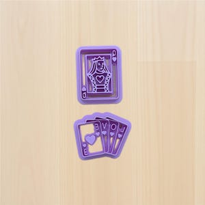 Könnte beinhalten: Lila Kunststoff-Ausstecher in Form von Spielkarten. Einer ist die Herzkönigin, der andere ein Satz von vier Karten mit dem Wort "LOVE" und einem Herzsymbol. Die Ausstecher liegen auf einer hellen Holzoberfläche.