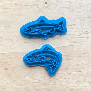 May include: Two blue plastic fish-shaped cookie cutters. The cutters are in the shape of a fish with detailed fins, gills, and eyes. The cutters are designed to cut out fish shapes from dough or other materials.