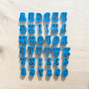 Puede incluir: Un conjunto de cortadores de galletas de plástico azul con letras y números. Las letras y los números están en negrita, con una fuente sans-serif. Los cortadores están dispuestos en filas sobre una superficie clara.
