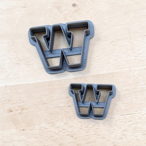 May include: Two gray cookie cutters in the shape of the capital letter W. The cutters feature a thick, raised border and a flat base, with one cutter noticeably larger than the other. Both items have a textured surface finish from the manufacturing process.