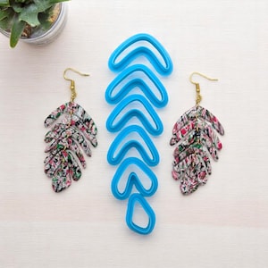 May include: A pair of leaf-shaped earrings with a colorful floral pattern, displayed next to blue polymer clay cutters. The earrings have gold-colored hooks. The cutters are in various sizes and shapes.