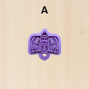 Puede incluir: Un cortador de galletas de pl&aacute;stico morado con forma de p&aacute;jaro estilizado. El cortador presenta detalles intrincados, incluyendo patrones florales y geom&eacute;tricos. La letra "A" est&aacute; impresa en negro en la parte superior de la imagen. El cortador est&aacute; sobre una superficie de madera clara.