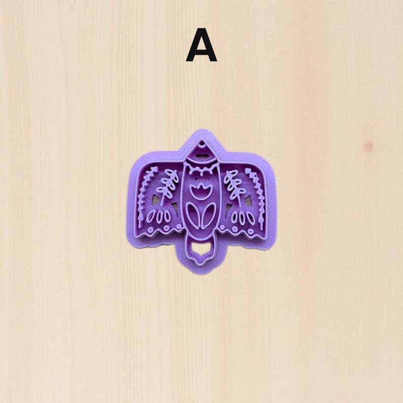 Puede incluir: Un cortador de galletas de pl&aacute;stico morado con forma de p&aacute;jaro estilizado. El cortador presenta detalles intrincados, incluyendo patrones florales y geom&eacute;tricos. La letra "A" est&aacute; impresa en negro en la parte superior de la imagen. El cortador est&aacute; sobre una superficie de madera clara.