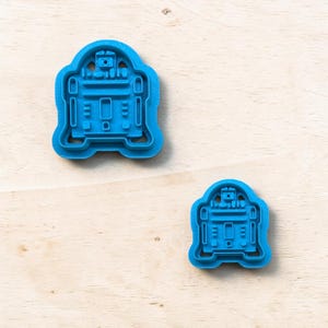 以下が含まれることがあります： ロボットの形をした2つの青いプラスチック製のクッキーカッター。カッターは、長方形と円形の要素を備えた詳細なデザインです。1つのカッターはもう一方よりも大きいです。