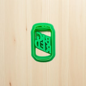 May include: A green plastic cookie cutter in the shape of a beer can. The word "BEER" is cut out of the center of the can. The cookie cutter is on a light wood surface.