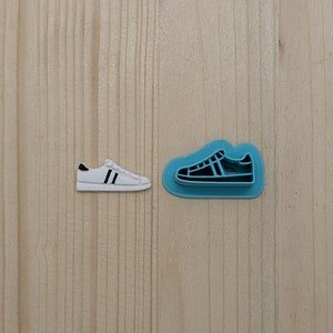 May include: A white trainer with black stripes and a matching shoe-shaped cookie cutter in light blue. The trainer has a white sole and a black heel. The cookie cutter is on a wooden surface.
