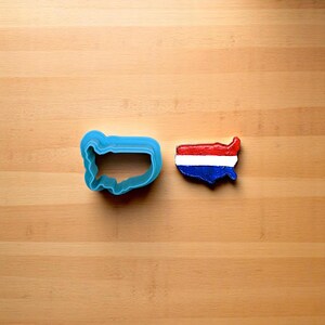 May include: A blue plastic cookie cutter shaped like the United States of America next to a red, white, and blue cookie in the shape of the United States.