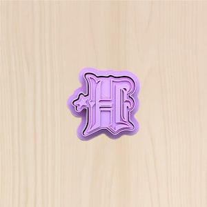May include: A lavender-colored cookie cutter in the shape of the letter "H". The letter has an ornate, stylized design with a star on the left side. The cutter is made of plastic and has a raised edge for easy use. The background is a light wood grain.