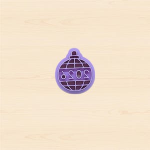 May include: A purple plastic cookie cutter in the shape of a disco ball with the word "SOSO" in the center. The cutter has a handle at the top and is set against a light wood grain background.