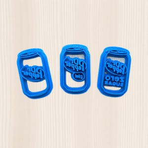 May include: Three blue plastic cookie cutters in the shape of soda cans. The cutters have the words "Dr Pepper" and "Diet" or "Zero Sugar" printed on them. The cutters are arranged on a light beige surface.