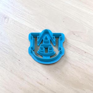 May include: A bright blue plastic cookie cutter shaped like the Auburn University AU logo. The cutter features a bold, stylized letter A with a U integrated into the center, designed with a thick frame for cutting dough into the specific collegiate emblem.