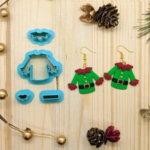 May include: A pair of green and red elf-themed earrings with gold hooks. The earrings are made of polymer clay and feature a green elf shirt with a black belt and red sleeves. The earrings are accompanied by a set of cookie cutters in the shape of an elf shirt, a holly leaf, and a rectangle.