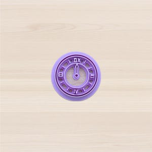 Può includere: Un tagliabiscotti in plastica viola a forma di orologio. Il quadrante dell'orologio ha numeri romani e lancette che indicano la mezzanotte. Il tagliabiscotti è rotondo con un bordo scanalato.
