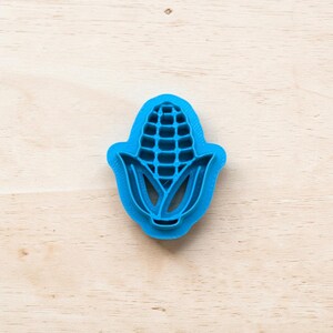 Peut inclure: Emporte-pièce bleu en forme d'épi de maïs. Le découpoir en plastique présente un motif détaillé de l'épi de maïs et de ses feuilles. Posé sur une surface en bois clair.