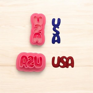 May include: Pink USA cookie cutters and a blue and red USA cookie. The cookie cutters are in the shape of the letters U, S, and A. The letters are also shown in blue and red.