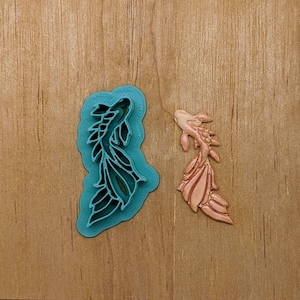Puede incluir: Dos cortadores de galletas con forma de pez, uno azul verdoso y otro rosa. El cortador azul verdoso es una silueta detallada de un pez con aletas y cola. El cortador rosa es un pez más estilizado con un cuerpo redondeado y una cola larga.