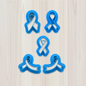 Puede incluir: Cinco cortadores de galletas con forma de lazo azul. Los cortadores tienen un contorno azul claro y un diseño de lazo blanco. Los cortadores están dispuestos sobre una superficie de vetas de madera clara.