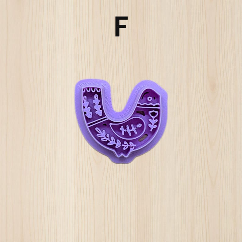 Puede incluir: Un cortador de galletas morado con forma de p&aacute;jaro, con patrones florales y geom&eacute;tricos decorativos. La letra "F" est&aacute; encima del cortador de galletas. El dise&ntilde;o del p&aacute;jaro incluye un ojo peque&ntilde;o, un pico y una cola.