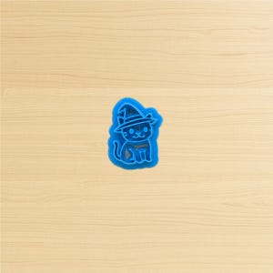 May include: A blue plastic cat-shaped cookie cutter wearing a witch hat. The cat is sitting, with a smiling face and a small collar. The cutter is on a light brown wooden surface.