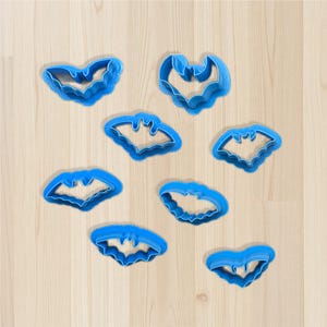 May include: A collection of eight blue plastic bat-shaped cookie cutters. The cutters are arranged on a light wood-grain surface. Each cutter has a detailed outline of a bat, perfect for Halloween or themed baking.