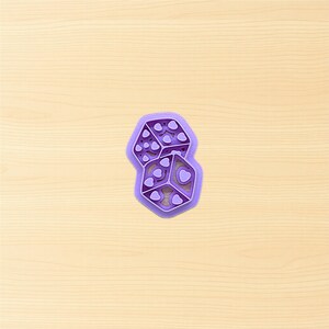 May include: A purple cookie cutter shaped like two dice. Each die features heart-shaped markings. The cutter is made of plastic and rests on a light brown wooden surface.