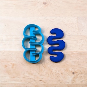Pode incluir: Um cortador de biscoitos de plástico azul-claro com um formato ondulado e serpentino, acompanhado por uma peça sólida azul-escura com o mesmo formato. O cortador possui uma barra de suporte vertical central que une as três alças onduladas.