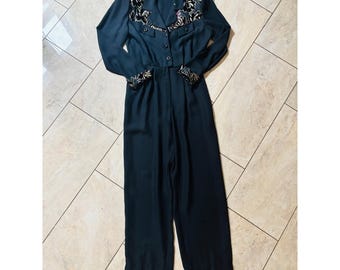 Könnte beinhalten: Ein schwarzer Jumpsuit mit langen Ärmeln, Knopfleiste, Kragen und dekorativen Einsätzen an Brust und Ärmelbündchen mit dunklem Blumenmuster. Das Kleidungsstück verfügt über ein tailliertes Oberteil und eine gerade geschnittene Hose.