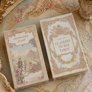 May include: Two rectangular card deck boxes with cream-colored backgrounds and ornate gold borders. The left box features a rider on a horse and the text Lenormand deck 36 cards. The right box displays cherubs and the text LE JARDIN DU ROI TAROT.