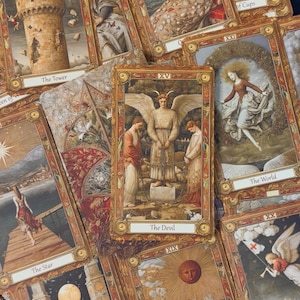 Puede incluir: Un conjunto de cartas del tarot con marcos dorados ornamentados. Las cartas presentan ilustraciones detalladas con títulos como "El Diablo", "La Torre" y "El Sol". La paleta de colores incluye tonos cálidos de marrón, dorado y rojo, creando una estética vintage.
