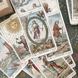 Op de afbeelding: Een verzameling tarotkaarten in vintage-stijl verspreid over een oppervlak. Zichtbare kaarten zijn onder meer De Wereld, De Maan, 8 Kelken en De Dwaas, met gedetailleerde illustraties van figuren, dieren en hemelse symbolen in gedempte tinten.